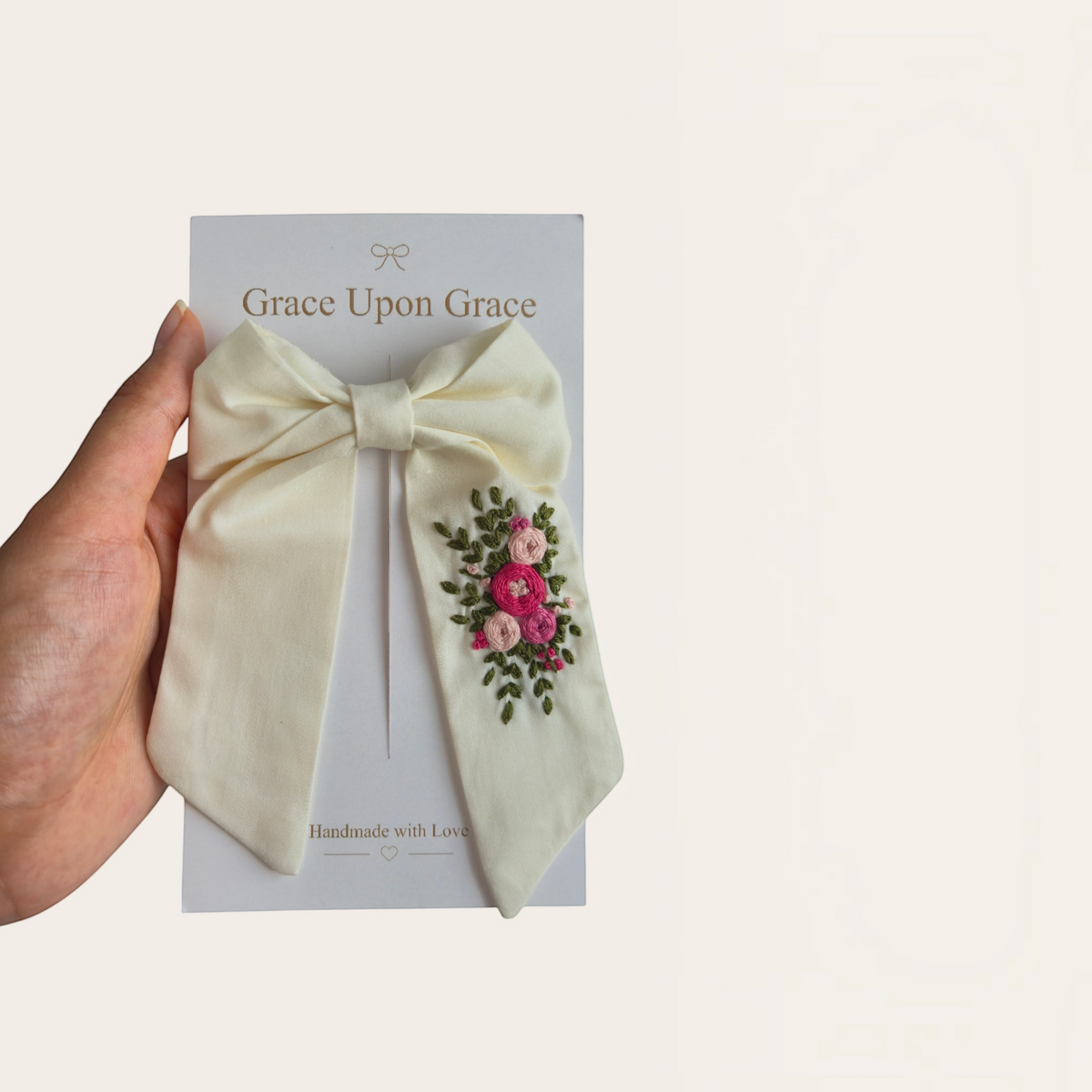 Grace Upon Grace ivory sailor bow featuring hand-embroidered pink roses and green leaves on a display card.
